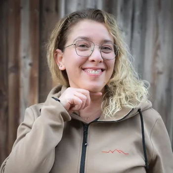 Eine lächelnde Frau mit Brille und lockigem Haar steht vor einer Holzwand und deutet auf ihr Kinn.