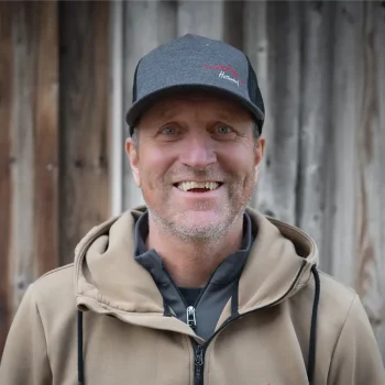 Ein lächelnder Mann mit hellbraunem Kapuzenpulli und grauer Mütze steht vor einer verwitterten Holzwand.