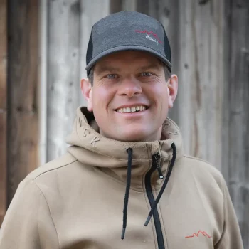 Mann in hellbraunem Kapuzenpulli und grauer Mütze, der vor einer Holzwand lächelt.