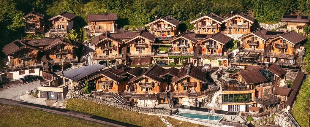 Luftaufnahme einer Gruppe von Holzchalets an einem Hang, umgeben von Grün und Bäumen.